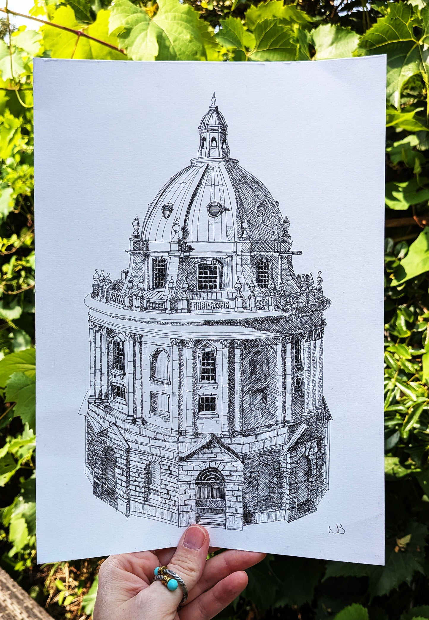Hand holding a black and white architectural drawing of a dome against a green leafy background