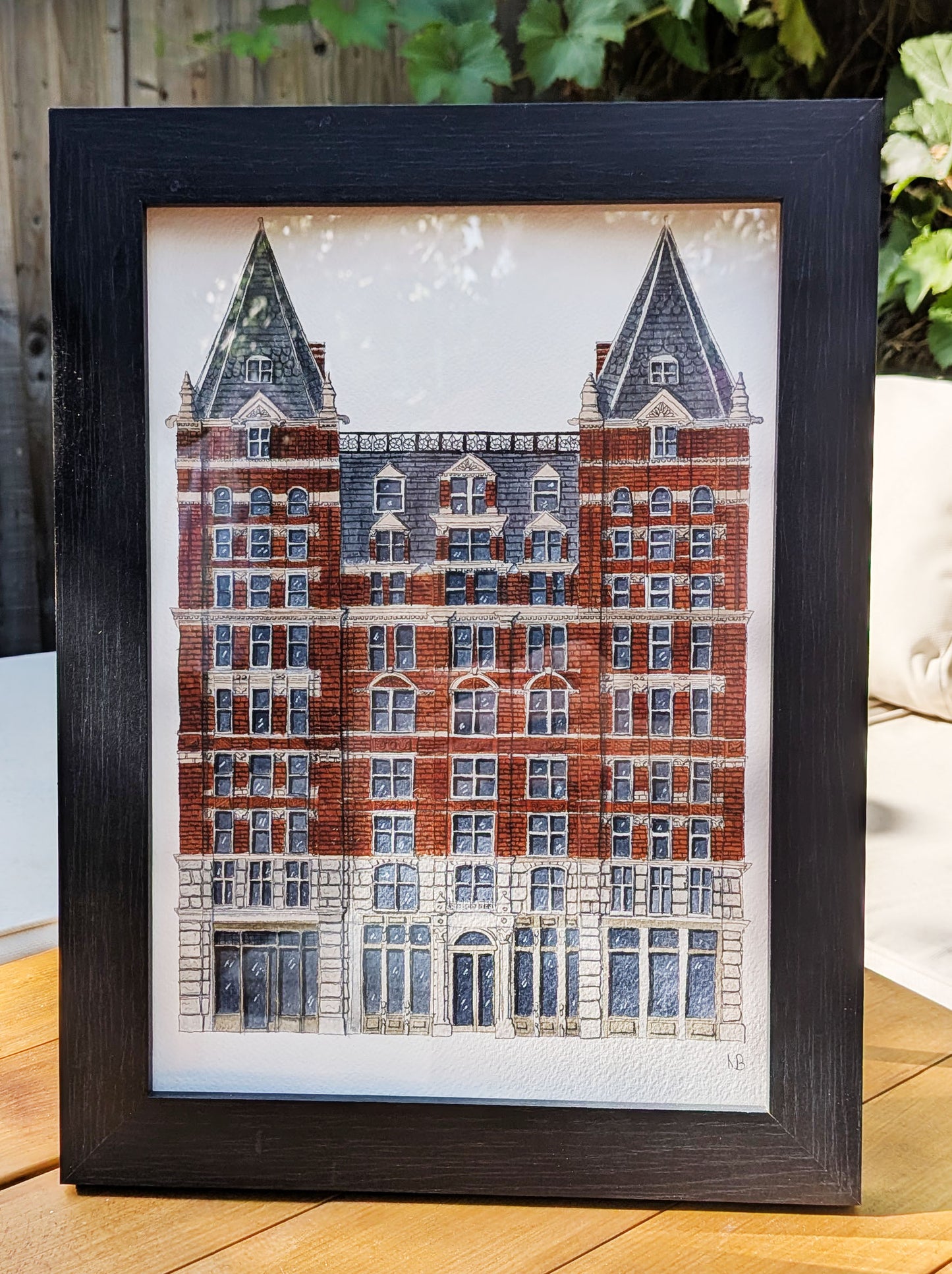 Framed artwork of a detailed building on a wooden surface with plants in the background