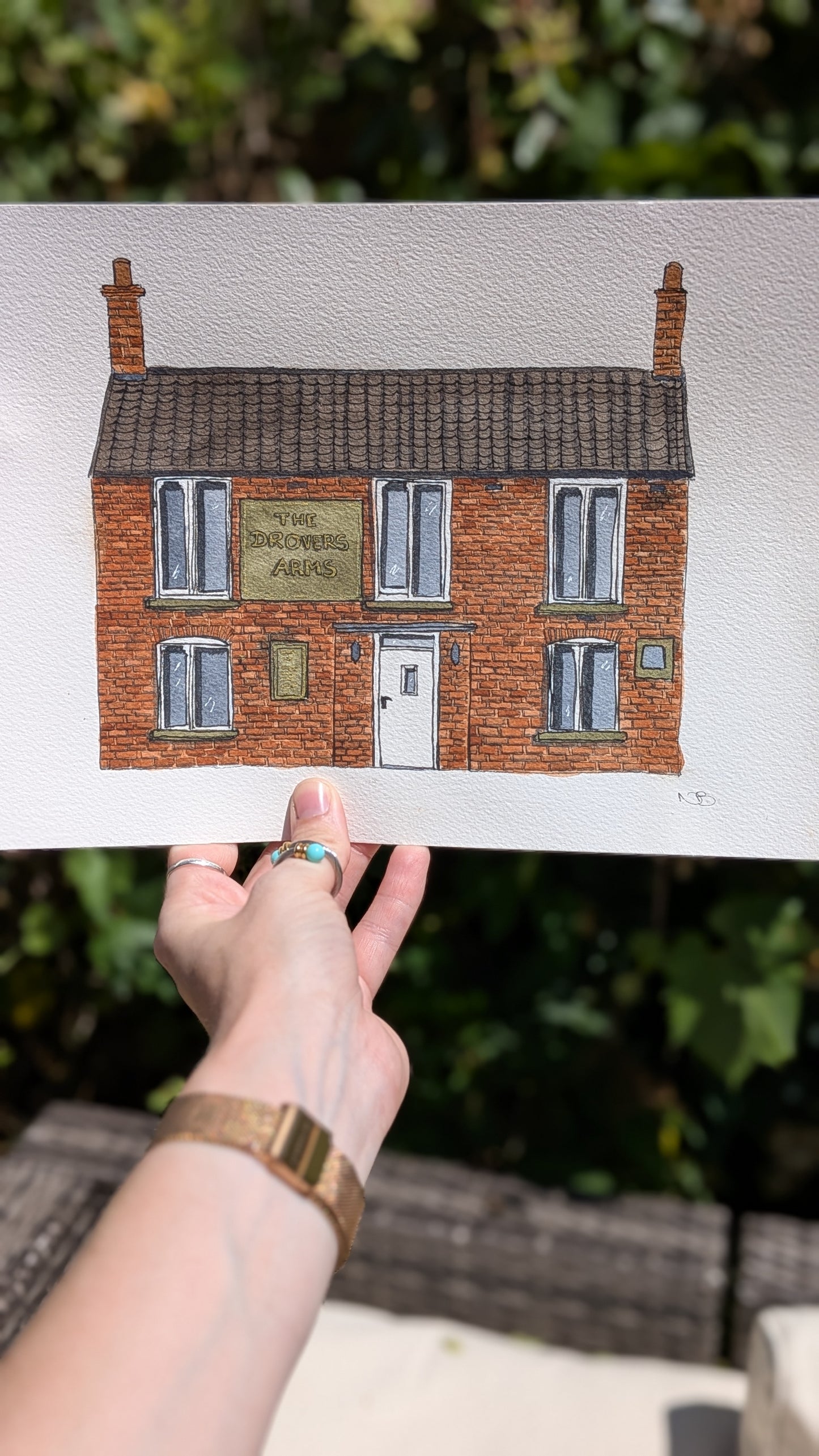 Hand holding a print of a brick building with a sign, against a blurred outdoor background.