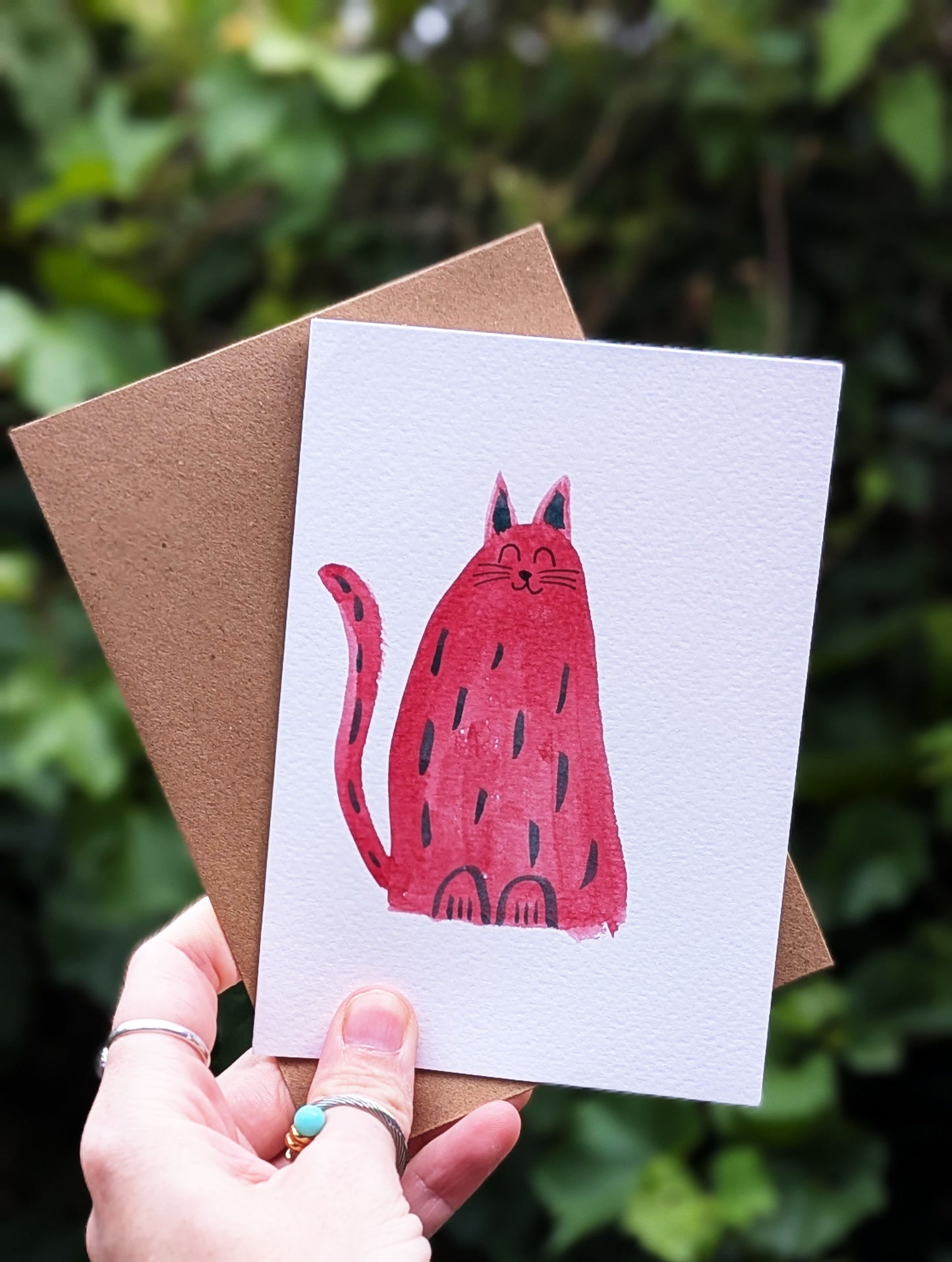 Card with a red cat illustration held by a person against a green leafy background