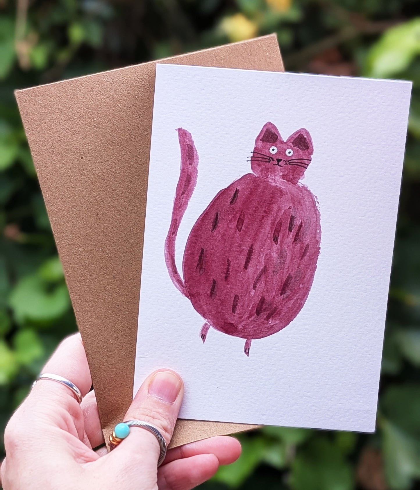Card with a pink cat illustration held by a hand against a green leafy background