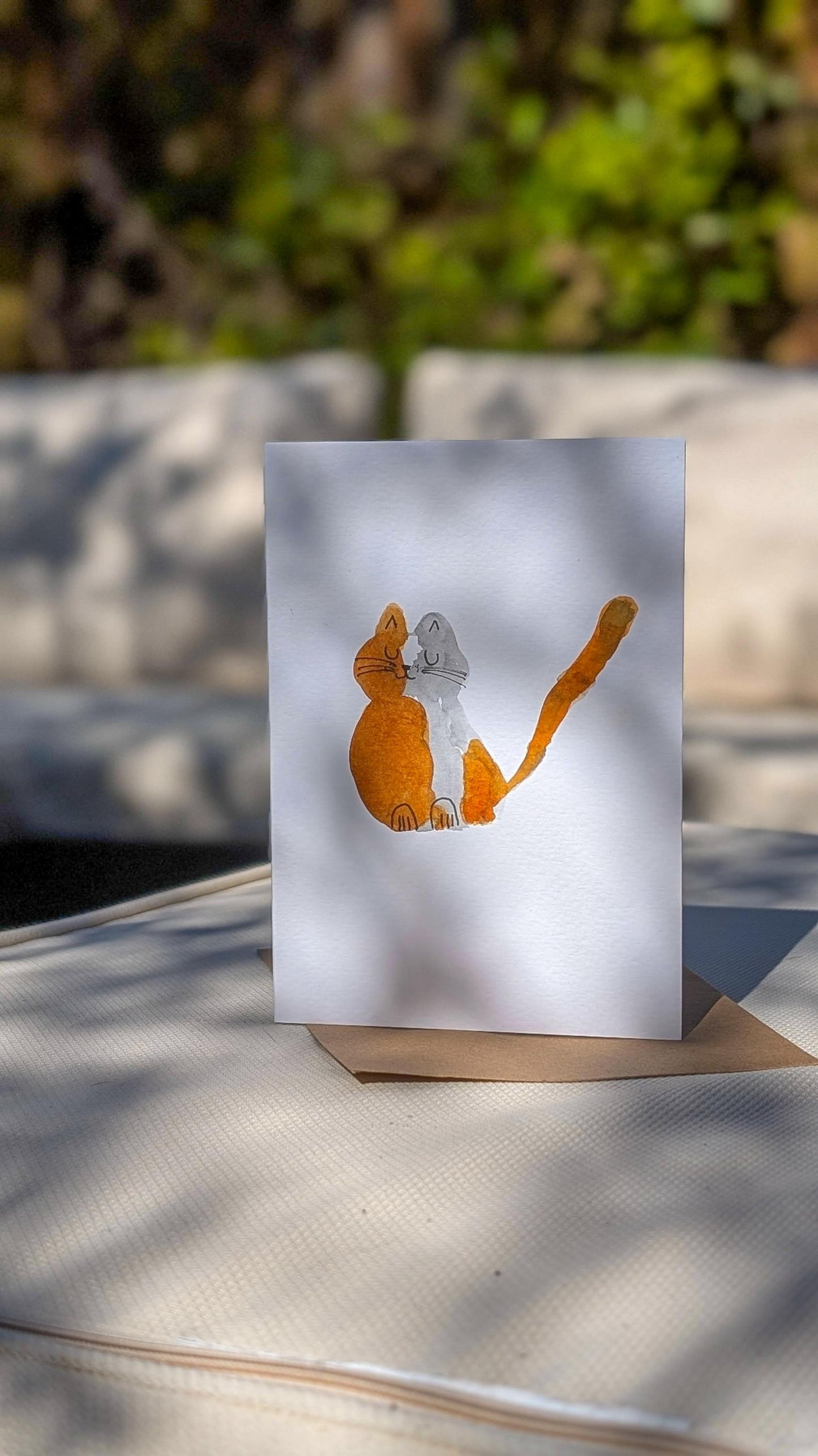 A hand-made watercolour greetings card featuring an abstract orange cat design on a white background, displayed on a table with a blurred natural setting.
