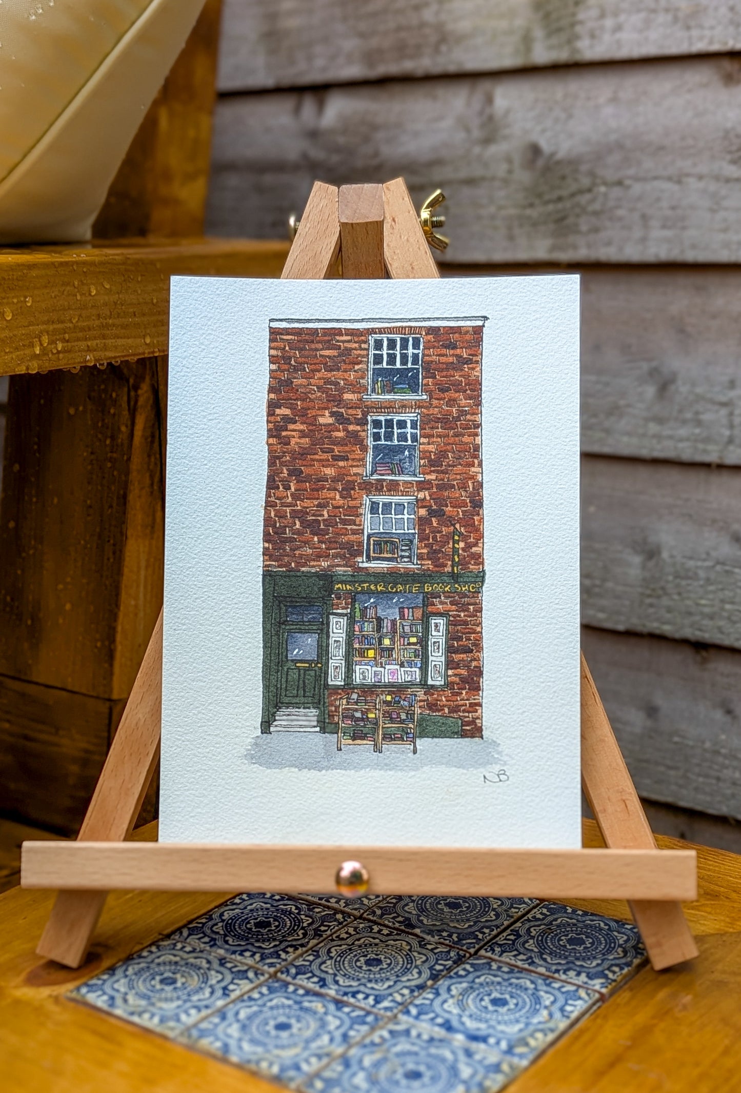Art print of a brick building on a white card with a wooden easel against a wooden background.