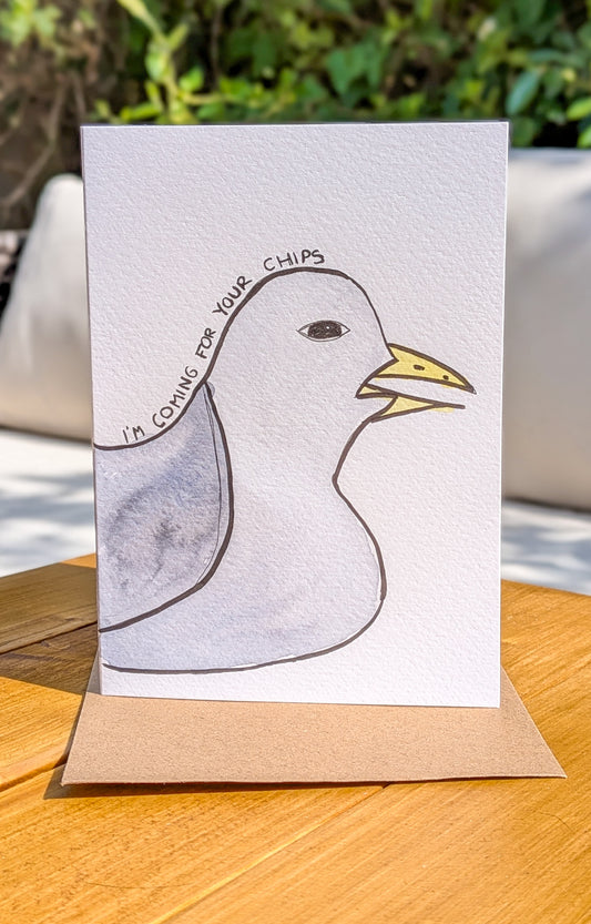 Hand-painted watercolor greetings card featuring a seagull with the text 'I'm coming for your chips' on the front, placed on a wooden surface with foliage in the background.