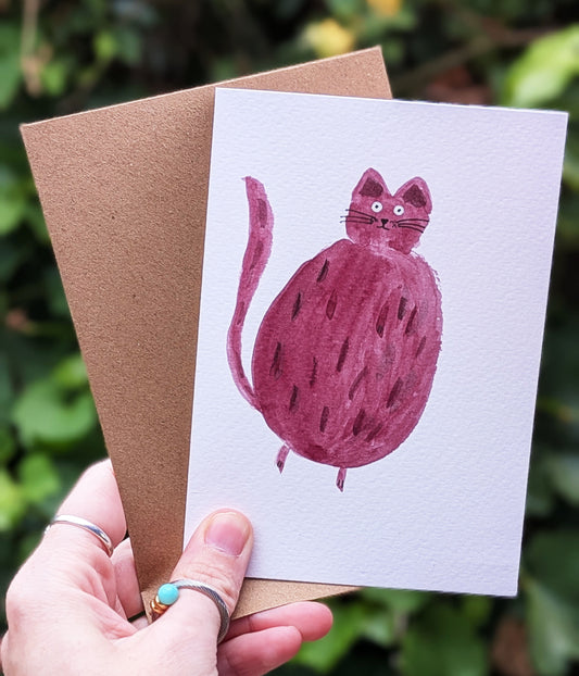 Card with a pink cat illustration held by a hand against a green leafy background