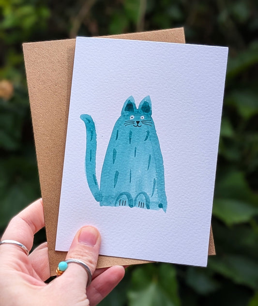 Card with a blue cat illustration held by a hand against a green leafy background
