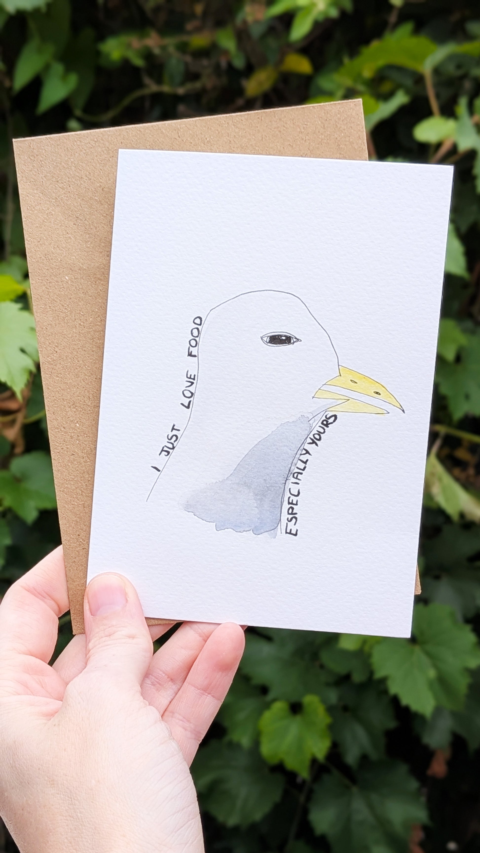 Hand holding a card with a seagull illustration and text against a green leafy background