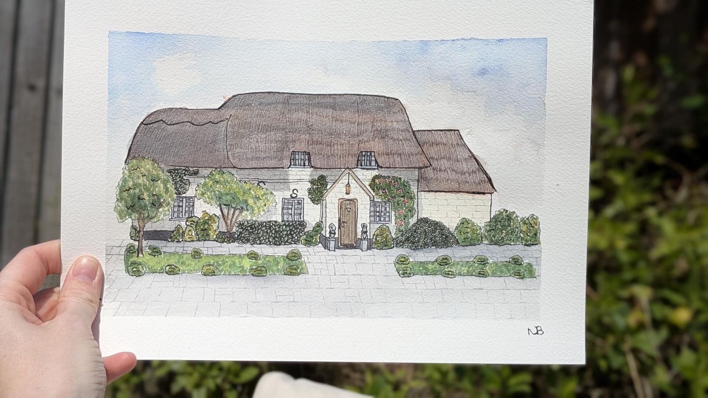 hand holding a painting of a thatched roof cottage