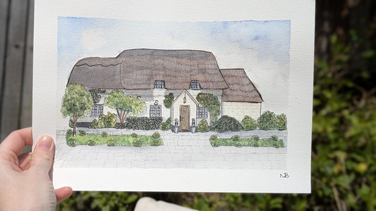 hand holding a painting of a thatched roof cottage
