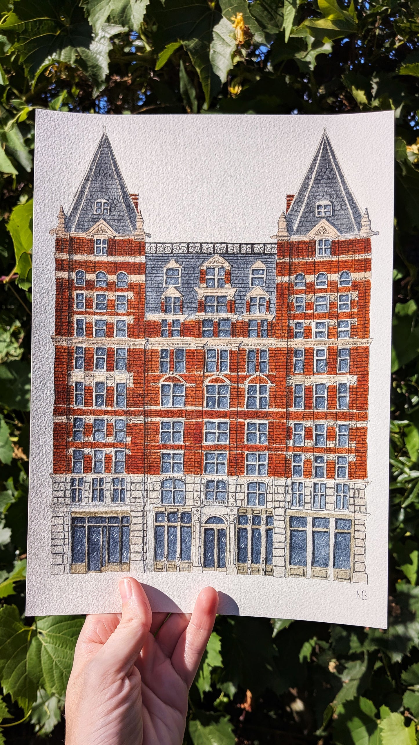 Hand holding a print of a building with red brick and blue accents against a green leafy background