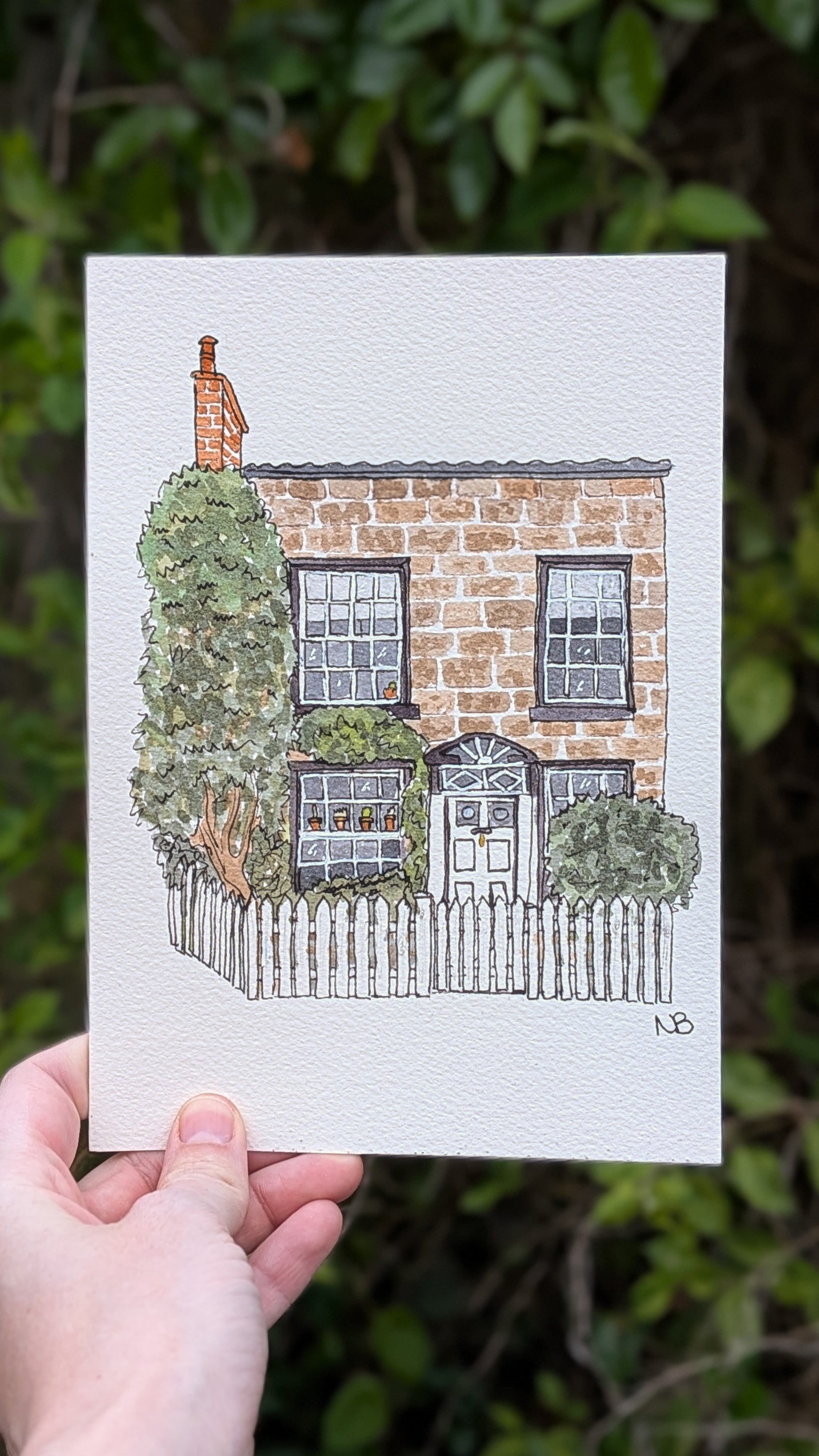 A framed hand-drawn watercolor illustration of a house on a wooden table, with greenery and a blue sky depicted on the building.