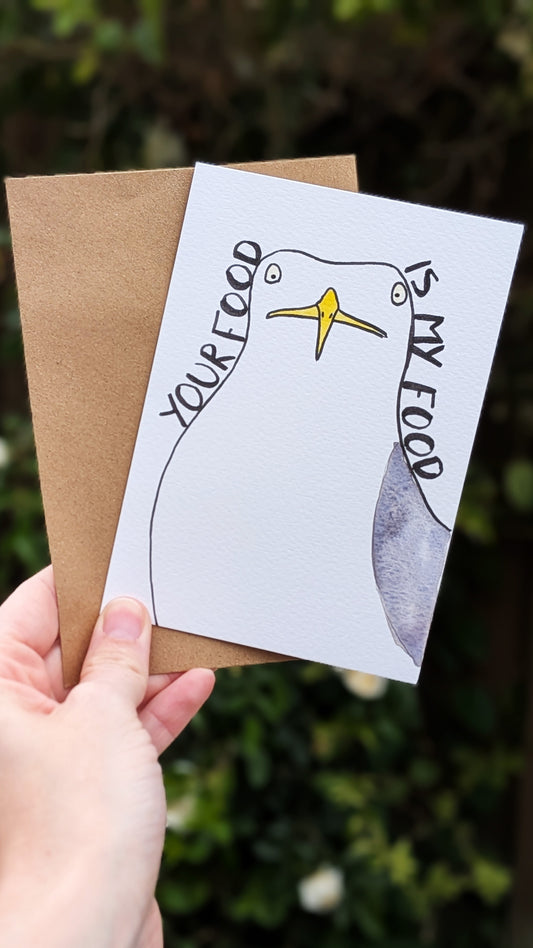 Hand holding a card with a humorous design of a bird and text, against a blurred natural background.